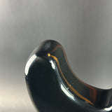 Close-up of a glossy black curved object, likely a decorative sculpture or vessel, with a small amber accent visible along the inner curve, photographed against a neutral gray background.