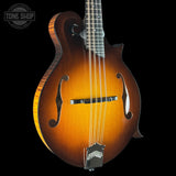 A close-up studio shot of an electric mandolin guitar against a solid black background. The mandolin features a classic sunburst finish transitioning from a dark brown outer edge to a bright amber-orange center, with silver metal hardware including a bridge and tailpiece.