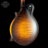 Close-up of the back of a mandolin with a glossy, figured wood finish showing warm amber and brown tones against a black background. A Tone Shop watermark is visible in the top-left corner.