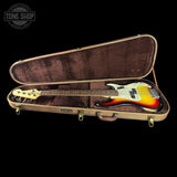 A Fender Stratocaster electric bass guitar in its open hard case, photographed against a solid black background. The bass features a classic sunburst finish with yellow, orange, and black gradients, a white pickguard, and gold hardware.