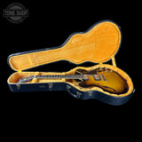 A sunburst electric guitar displayed inside an open black hard case with a gold-foiled velvet interior, set against a solid black background. A Tone Shop logo is visible in the top-left corner.