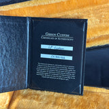 An open black Gibson Custom Certificate of Authenticity booklet showing handwritten identification labels for 'LP Custom' and serial 'CS300466', placed on a gold textured surface.