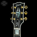 Close-up of the headstock of a Gibson Les Paul Custom electric guitar against a solid black background. The headstock is black with gold tuning pegs, white Gibson branding, and a mother-of-pearl inlay.