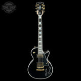 A black electric guitar with gold hardware and white binding, shown vertically against a solid black background. A circular 'TONE SHOP' watermark is visible in the top-left corner.