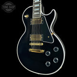 A black electric guitar with gold hardware and white binding, shown from a high angle against a black background. A Tone Shop watermark is visible in the top-left corner.