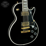 A close-up of a black electric guitar body with gold hardware, white binding, and a black pickguard, shown against a black background with a Tone Shop watermark in the top left.
