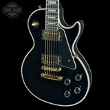 A black electric guitar with gold hardware and white binding, shown from a high angle against a black background. A Tone Shop watermark is visible in the top left corner.