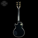 A black Gibson Les Paul Custom electric guitar shown from the back, featuring gold tuning pegs and a gold circular badge on the body, set against a solid black background with a Tone Shop watermark in the top left corner.
