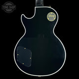 The back of a black Gibson electric guitar with a white binding and a gold Gibson logo sticker on the upper right side, shown against a solid black background.