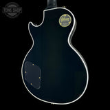 The back of a Gibson Les Paul Custom electric guitar in glossy black, featuring white binding, gold hardware, and a gold Gibson logo badge on the upper back.