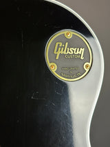 Close-up of a black guitar body with a circular gold Gibson Custom badge in the center, showing the brand name and 'Hand Crafted in Nashville, TN' text.