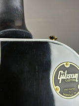 Close-up of the back of a black Gibson Custom electric guitar body, showing the gold and black Gibson Custom emblem and part of the neck.