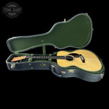 An acoustic guitar resting inside an open hard case on a black background, with a Tone Shop watermark in the top left corner.