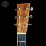 Close-up of the headstock of an acoustic guitar with a warm brown wood finish, six silver tuning pegs, and six strings, shown against a solid black background with a Tone Shop watermark in the top left corner.