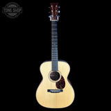 A natural wood acoustic guitar with a cutaway body, dark fretboard, and decorative pickguard, shown against a solid black background with a Tone Shop logo in the top-left corner.