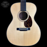 A front-facing acoustic guitar with a natural wood finish and dark pickguard, shown against a solid black background with a Tone Shop watermark in the top-left corner.