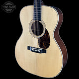 A close-up studio shot of an acoustic guitar body and upper neck against a solid black background. The guitar has a natural light wood top, dark brown sides, a dark tortoiseshell pickguard, and a black bridge.