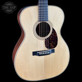 An acoustic guitar with a natural wood top, dark brown sides, and a tortoiseshell pickguard, shown against a solid black background with a Tone Shop watermark in the top-left corner.