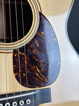 Close-up of an acoustic guitar body showing the glossy tortoiseshell-style pickguard, sound hole, strings, and light natural wood grain.