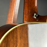 Close-up detail of an acoustic guitar body and neck, showing a glossy dark brown wood finish, a gold-toned edge trim, and part of the headstock against a neutral gray background.