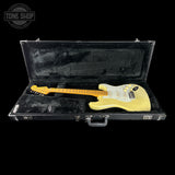 A yellow electric guitar with a white pickguard resting inside an open black hard case with black velvet lining, shown against a solid black background. A Tone Shop watermark is visible in the top-left corner.