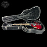 A red and black electric guitar with gold trim resting inside an open black hard case on a black background. A Tone Shop logo watermark is visible in the top-left corner.