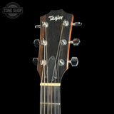 Close-up of the headstock of a Taylor acoustic guitar against a solid black background, showing the brand name 'Taylor' in white script on a dark wood headstock with six silver tuning pegs and strings.