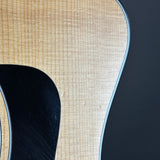 Close-up detail of an acoustic guitar body showing the natural wood grain top, a black soundhole surround, and a dark background.