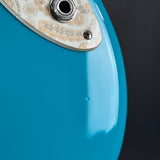 Close-up of the lower body of a turquoise blue electric guitar, showing part of the decorative white and gold pickguard and a metal jack connector against a dark background.
