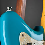 Close-up of the rear body of an electric guitar with a glossy turquoise-blue finish, a cream paisley pickguard, and part of a natural wood neck visible against a dark gray background.