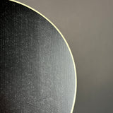 Close-up of a circular textured black product, likely a guitar headstock or decorative panel, with a light cream edge, set against a solid dark gray background.