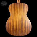 The back of a wooden acoustic guitar, showing a rich warm brown wood grain finish, photographed against a solid black background with a Tone Shop watermark in the top left corner.