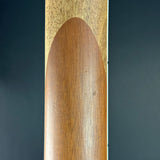 Close-up of a wooden object, likely a guitar headstock or decorative wood panel, showing a warm medium-brown arched inlay against a dark gray background.
