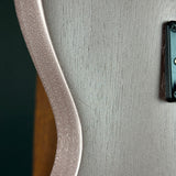 Close-up detail of a guitar body showing a light silver-gray wood grain finish, a curved edge, and part of a black hardware component.
