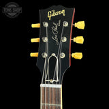 Close-up of the headstock of a Gibson Les Paul acoustic guitar, featuring a black headstock with gold Gibson branding, Les Paul script, and bright yellow tuning pegs against a black background.