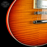 Close-up of a sunburst electric guitar body showing the flamed maple wood grain, white binding, chrome bridge, and part of the gold-toned pickup hardware against a black background.