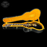 A black electric guitar inside an open hard case with a bright yellow-orange velvet interior, photographed on a black background. A Tone Shop watermark appears in the upper left corner.