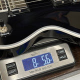 A close-up of a black electric guitar resting on a digital scale, showing the scale's blue LCD display reading 8.56 lbs.
