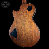 Close-up of the back of a wooden electric guitar body with a warm brown finish, black pickguard, and black circular control detail, shown against a black background. A Tone Shop watermark is visible in the upper left.