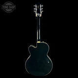 The back view of a glossy black electric guitar against a solid black background, featuring a circular Tone Shop watermark in the upper left corner.