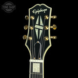 Close-up of the headstock of an Epiphone electric guitar against a solid black background, showing the gold tuning pegs, white brand lettering, and mother-of-pearl style inlays.