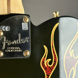 Close-up of the back of a Fender Corona electric guitar body, showing the black glossy finish, gold hardware, a gold and red flame graphic, and the Fender logo on the tailpiece.