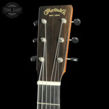 Close-up of the headstock of a Martin & Co acoustic guitar against a black background, showing the dark wood finish, gold brand lettering, and tuning pegs.