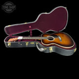 An acoustic guitar with a sunburst finish resting inside an open hard case with a burgundy velvet interior, shown against a solid black background. A Tone Shop logo is visible in the top-left corner.