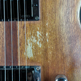 Close-up of a wooden electric guitar body showing a worn, chipped finish around the pickups and strings, with part of a chrome control visible at the bottom right.