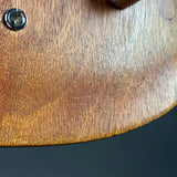 Close-up of a wooden guitar body showing the wood grain, a metal tuning peg hole, and a few visible scratches on the surface, with a dark background below.