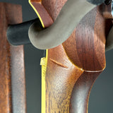Close-up of a polished brown wooden rifle stock with a black ribbed stock rest and a yellow tag attached, set against a dark gray background.