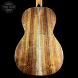 The back of a wooden acoustic guitar, showcasing a rich grain pattern in warm brown and golden tones, set against a solid black background with a Tone Shop watermark in the top left corner.