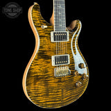 A close-up of a double-cutaway electric guitar body with a bright yellow-orange flamed maple top, gold hardware, and a white pickguard, shown against a black background with a Tone Shop watermark in the top left.