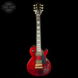 A Gibson Les Paul Custom electric guitar with a red flame finish, shown against a solid black background. The guitar features gold hardware, a black pickguard, and a dark fretboard with gold block inlays.